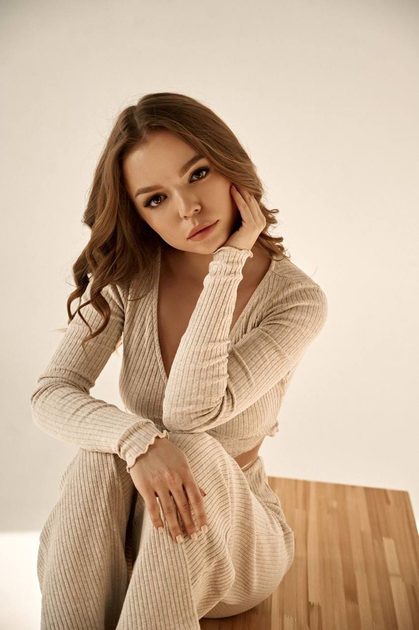 Giselle sits on wood in a beige ribbed knit set, hand on cheek, against a plain light background.