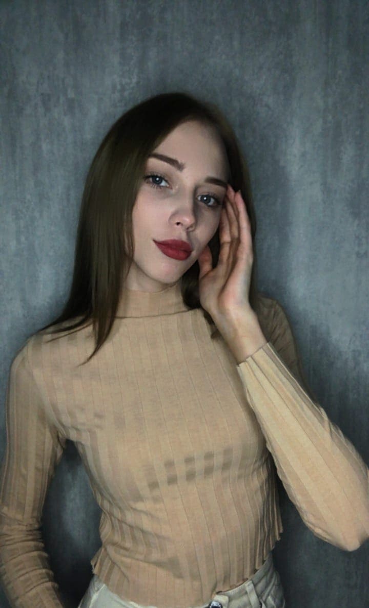 Dasha with long brown hair and red lipstick poses against a gray textured wall in a beige ribbed turtleneck, hand raised to her cheek.