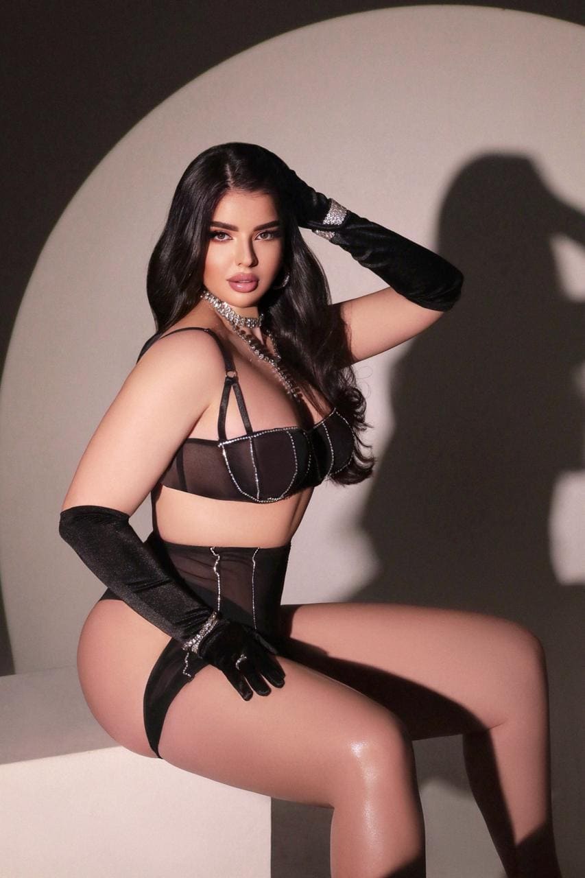 A woman sits on a white block in a studio, wearing black sheer lingerie, velvet gloves, and a choker, with one hand raised.