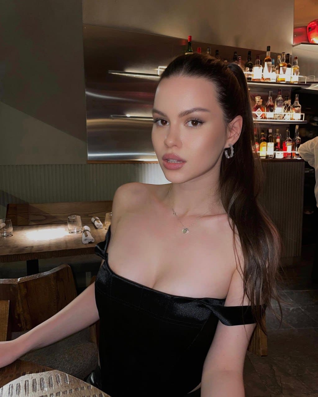 A woman with a high ponytail sits at a bar table in a black off-the-shoulder top, with shelves of liquor bottles visible behind her.
