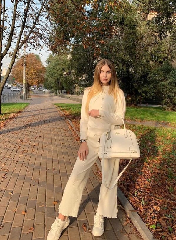 A woman stands on a brick sidewalk in an autumn park, wearing a cream long-sleeve top, ribbed trousers, and white sneakers, holding a white handbag.