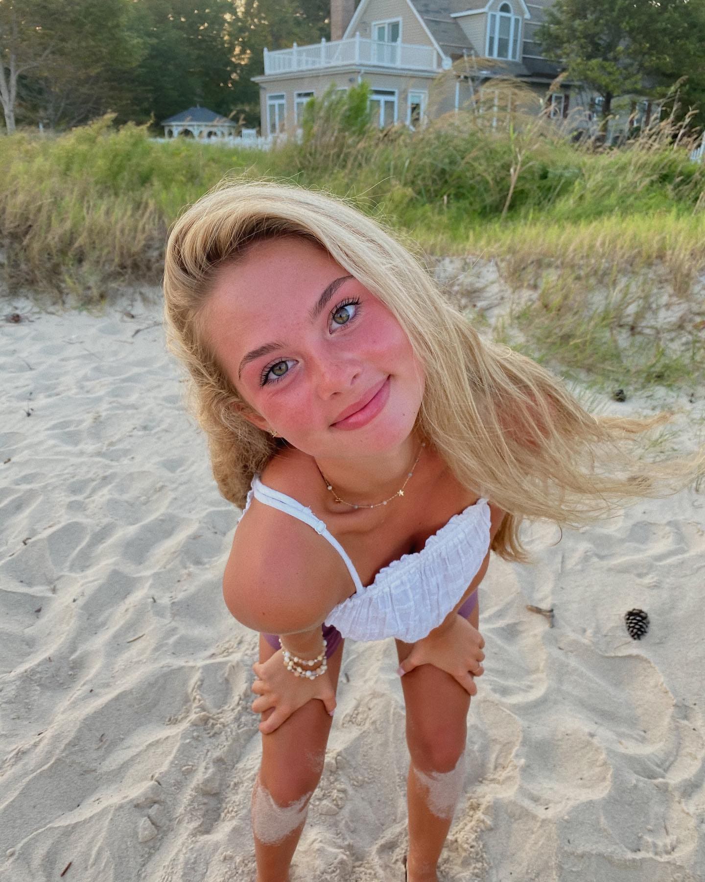 Vanessa leans forward on a sandy beach in a white ruffled bikini top and purple bottoms, with a house and dune grass behind her.