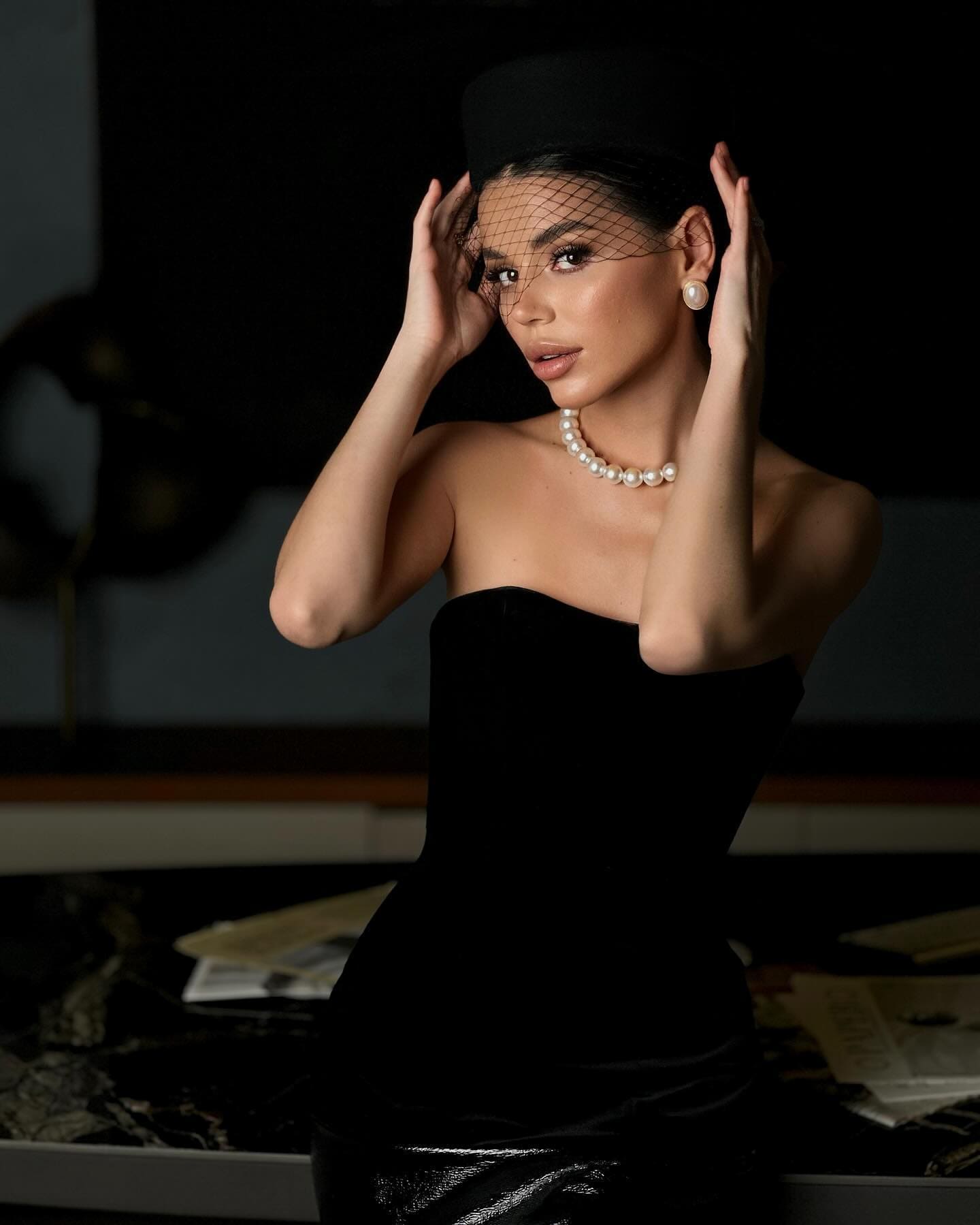 A woman in a black velvet dress and netted hat holds her head, wearing pearls, in a dim room with books.