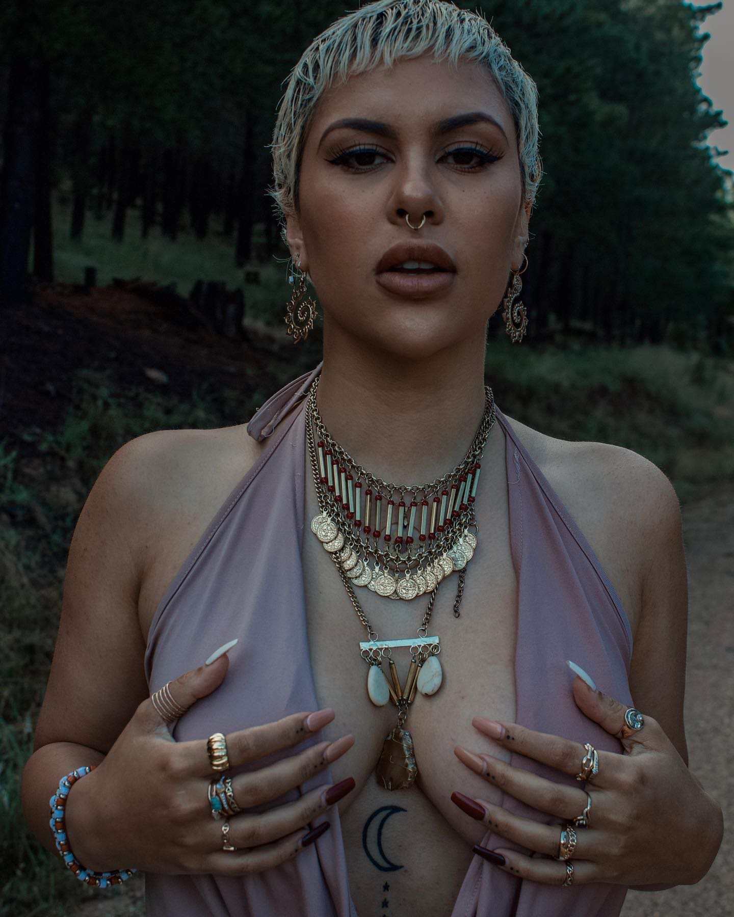 A person with short bleached hair stands outdoors in a dusty-pink halter top and layered necklaces, hands resting on their chest against trees.