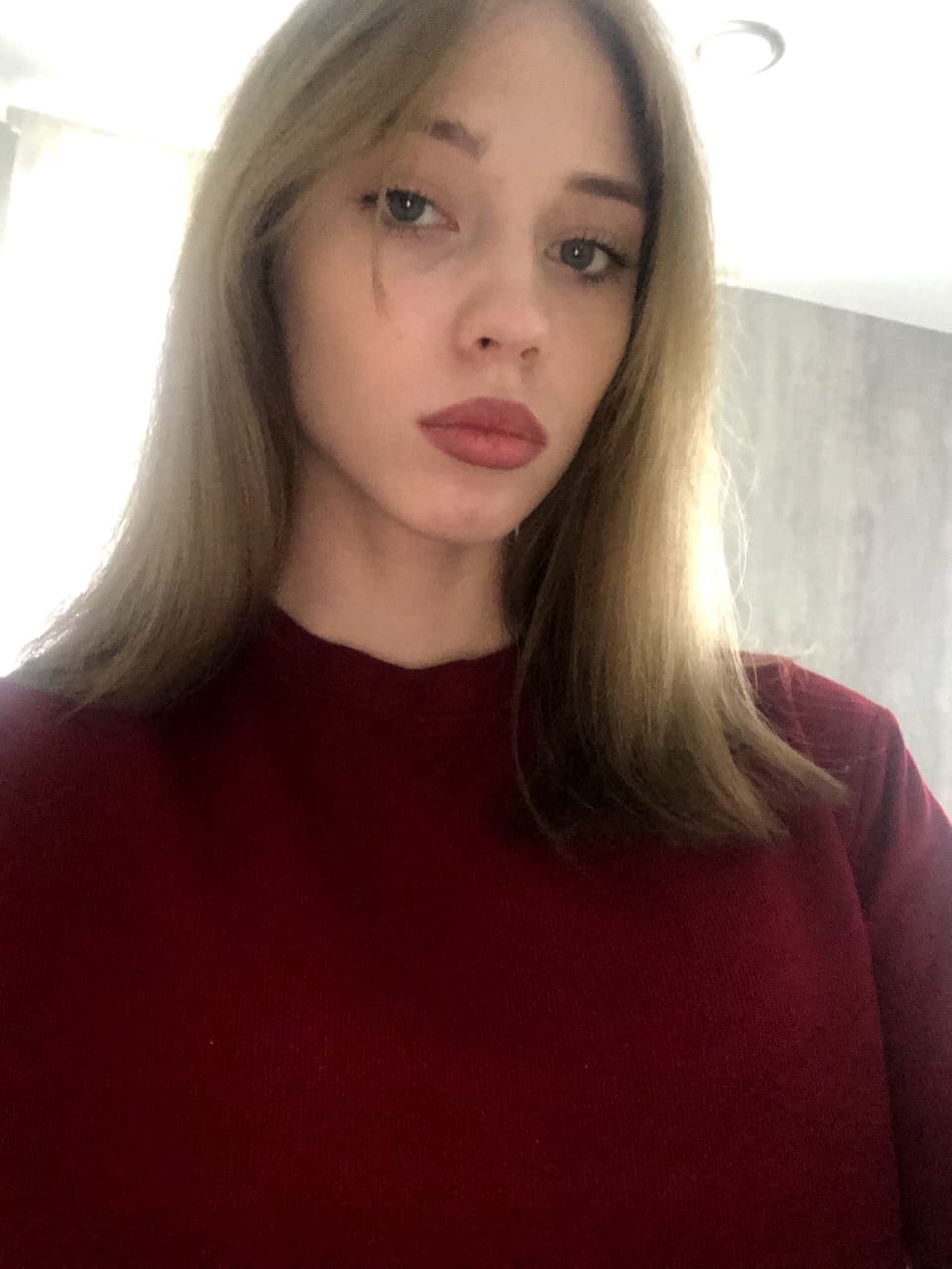 A close-up of a blonde woman with bangs wearing a dark red high-necked sweater, standing indoors near a bright window against a plain wall.