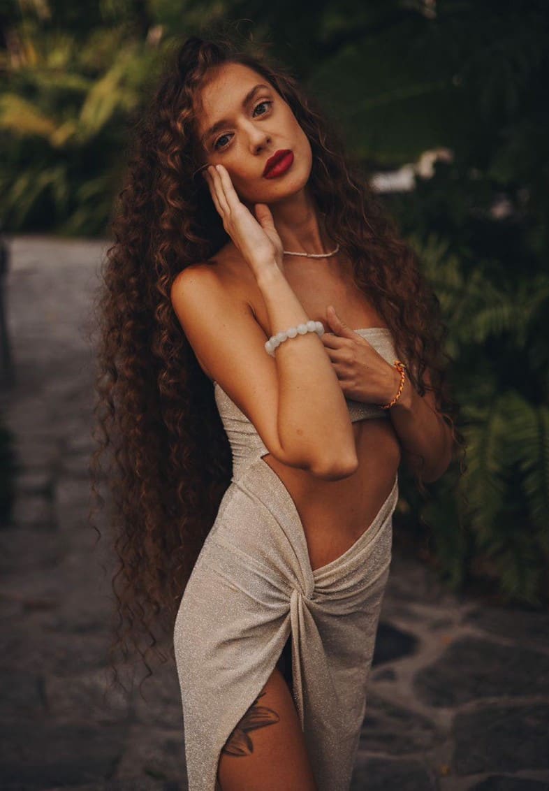 Montana stands on a stone path outdoors, wearing a strapless beige dress, with one hand on her cheek and the other on her chest.