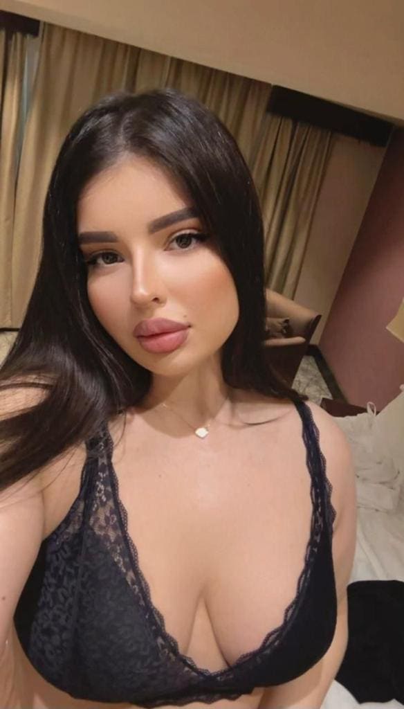 A woman poses in a black lace bralette and thin necklace in an indoor room with curtains and a bed in the background.