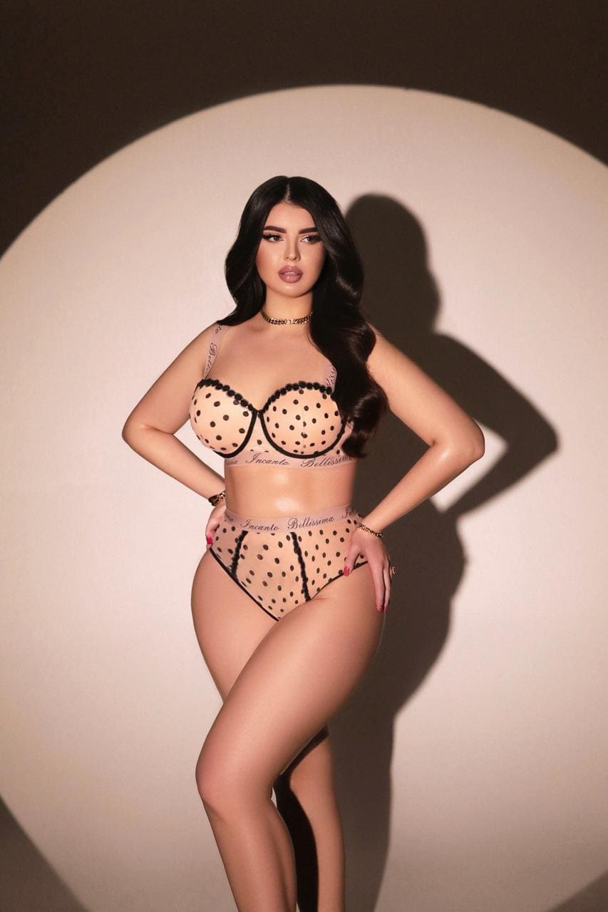A woman stands with hands on hips in a studio, wearing a sheer polka-dot lingerie set with black lace trim and a choker.