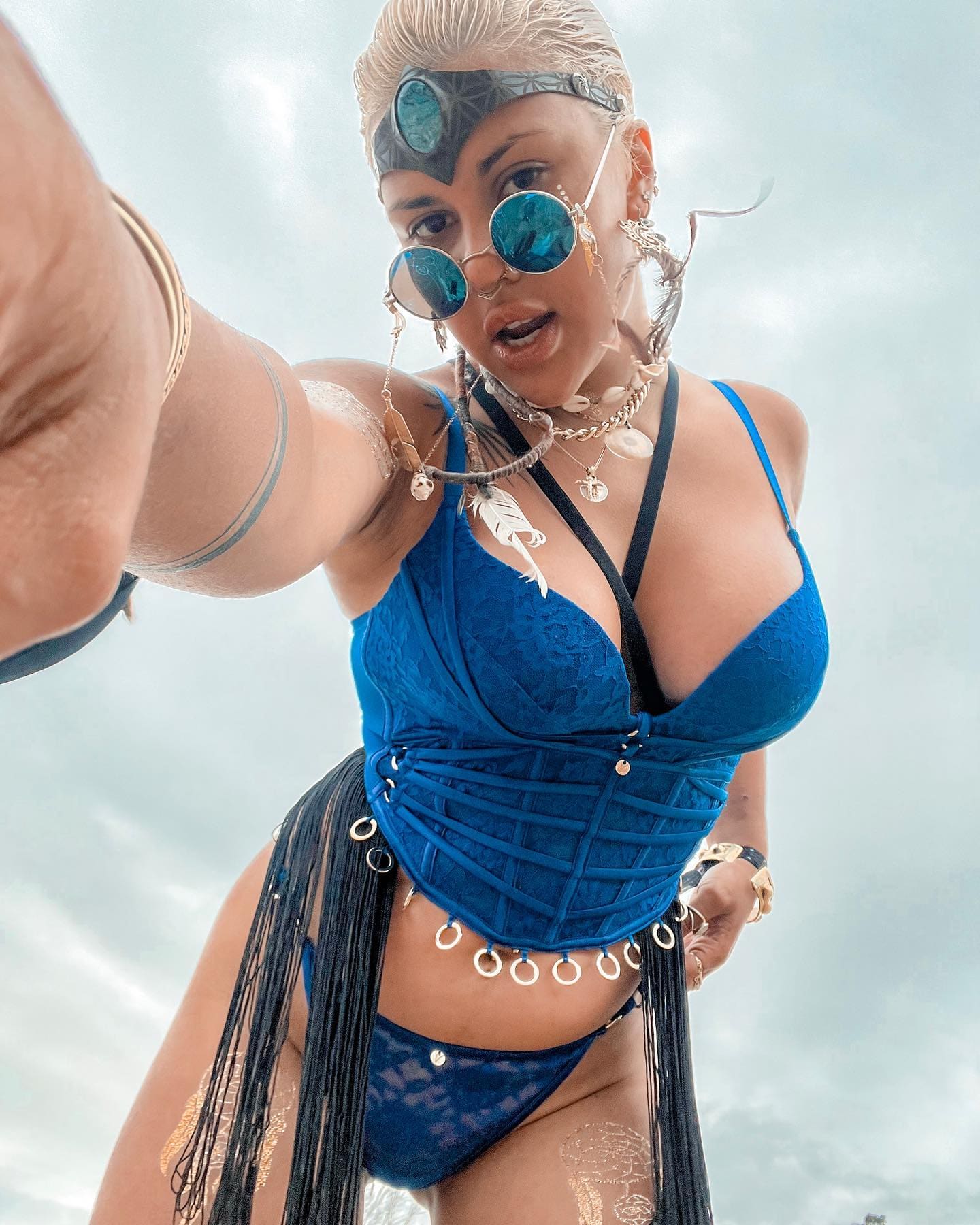 A selfie shows a person with wet blonde hair in a blue lace swimsuit and round sunglasses, posing against a cloudy sky background.