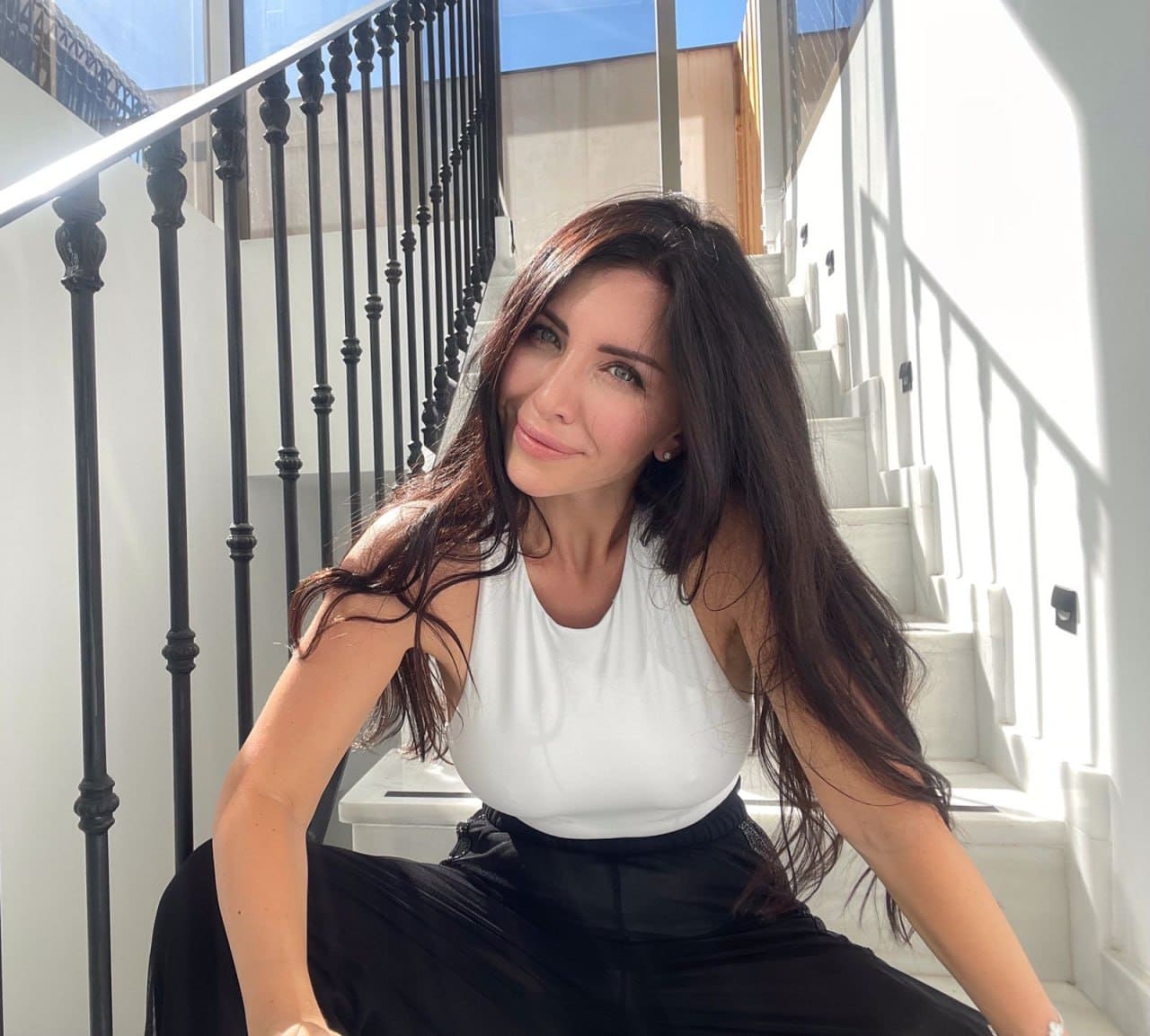 A woman sits on white stone steps beside a black railing, wearing a white sleeveless top and black pants, with sunlight casting shadows on the wall.