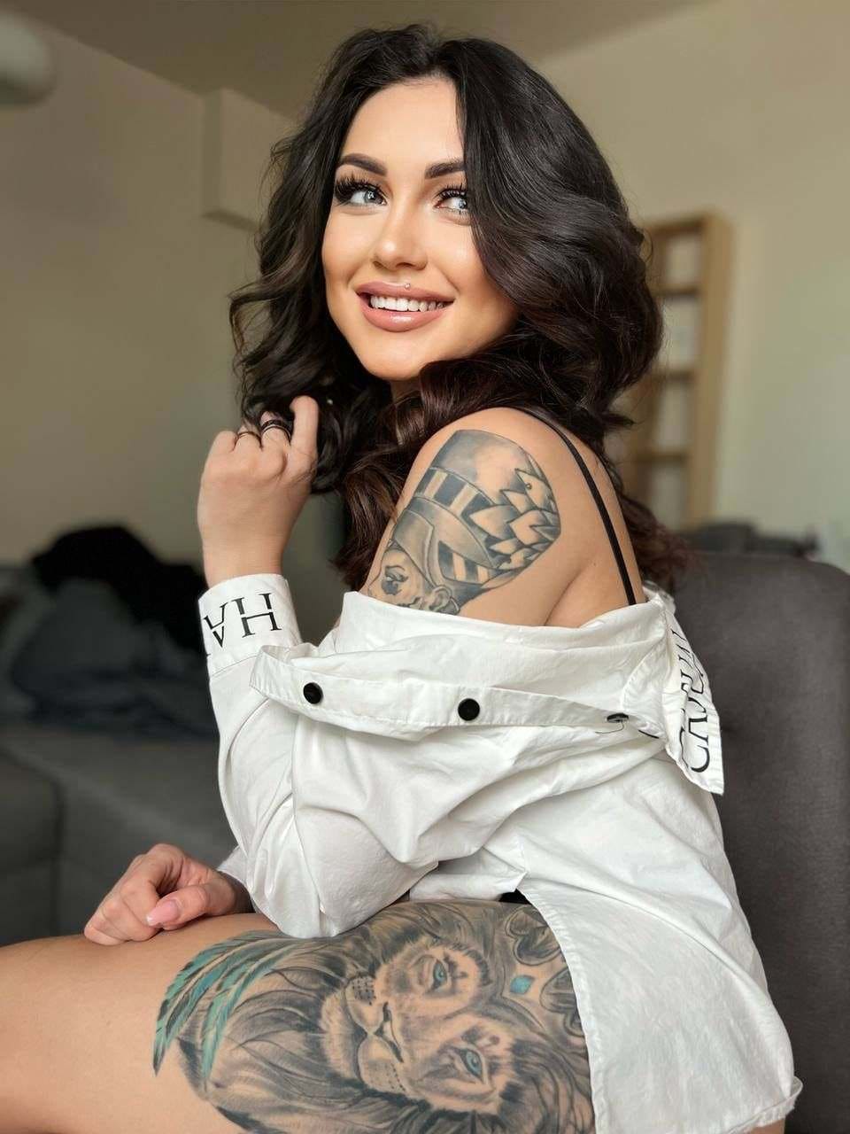 A woman sits on a gray couch in a white off-the-shoulder shirt, revealing arm and thigh tattoos.