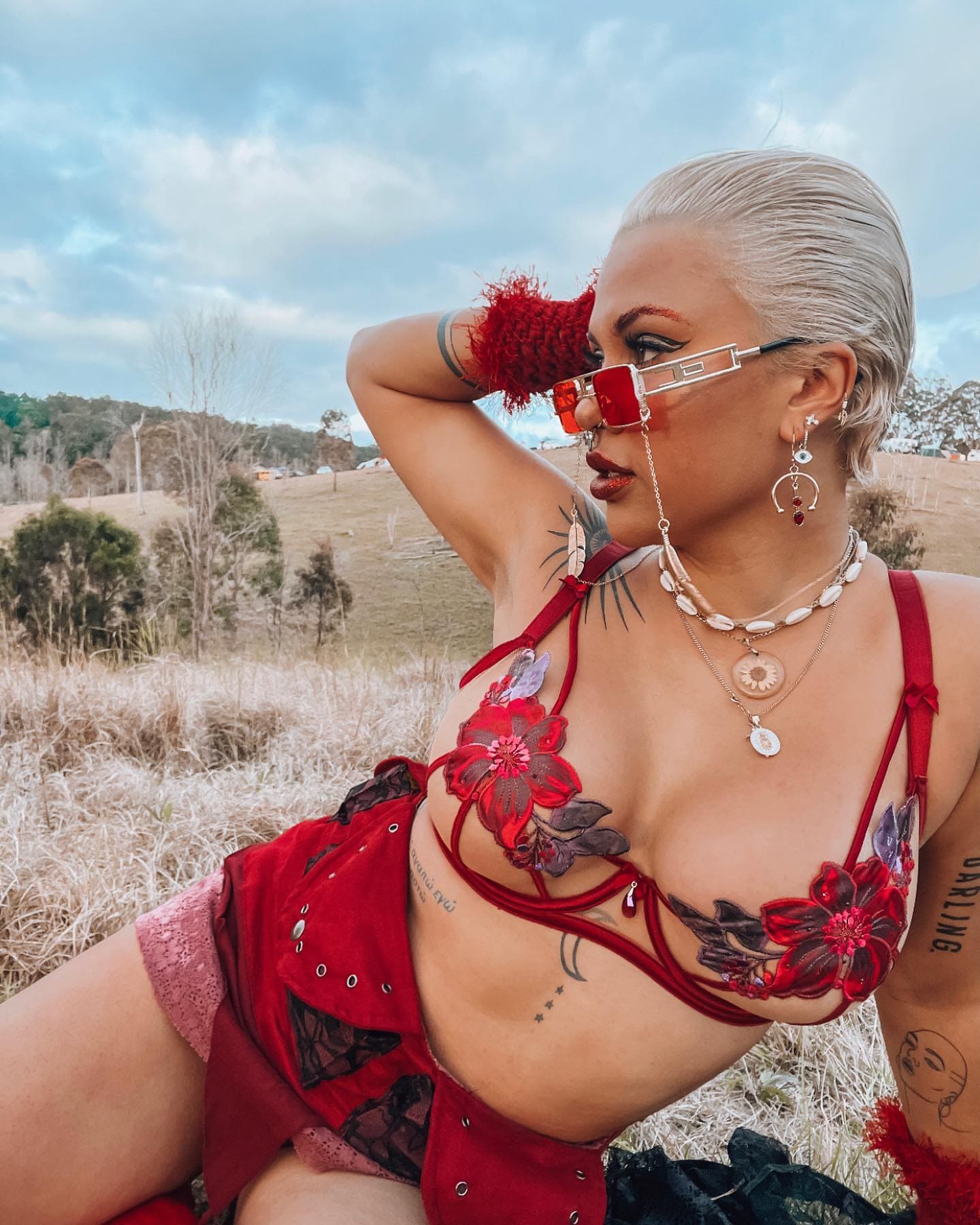 A person with platinum hair sits in a dry field wearing a red floral bikini top, red shorts, sunglasses, and a wrist cuff, one arm raised.