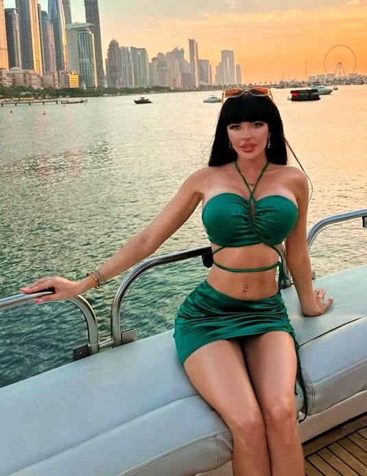 A woman sits on a boat bench in a green two-piece outfit, with sunglasses on her head, against a city skyline and water at sunset.