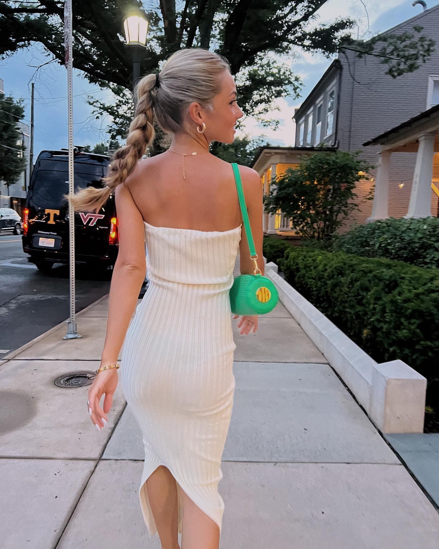 Seen from behind, a woman with a braided ponytail wears a white strapless dress and carries a green handbag while walking on a sidewalk.