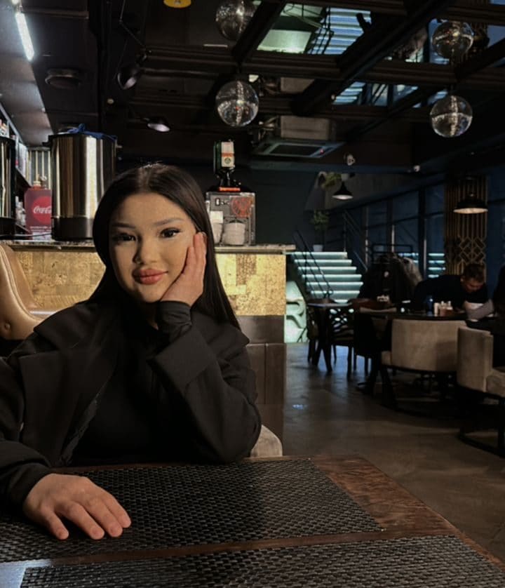 Karla sits at a table in a dimly lit restaurant, wearing a black jacket and resting her chin on her hand while looking toward the camera.