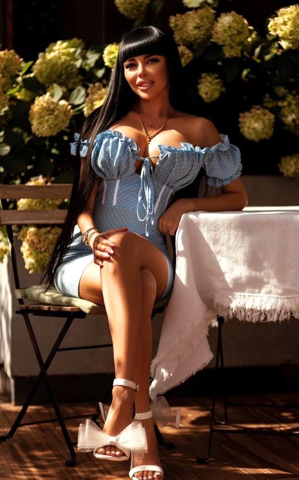 Melissa sits on a wooden chair in a blue polka-dot bodysuit and white bow sandals, against a backdrop of green foliage and white flowers.