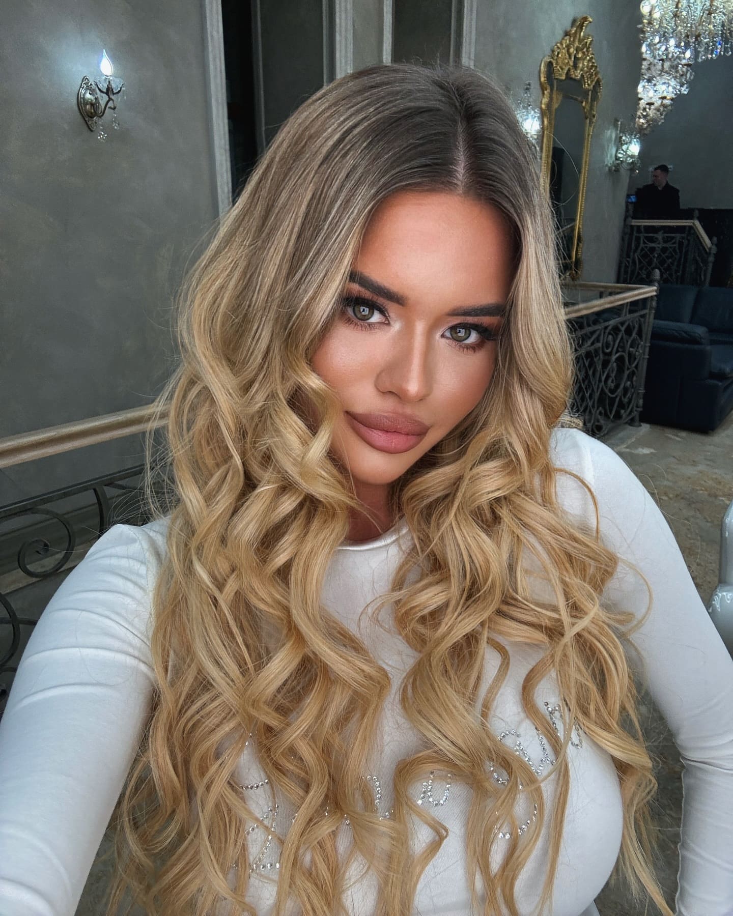 A woman with wavy blonde hair takes a selfie indoors wearing a white long-sleeved top, with a chandelier and gold mirror behind her.