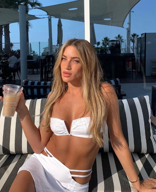 A woman in a white bikini top and wrap skirt sits on a striped lounge chair by a poolside bar.