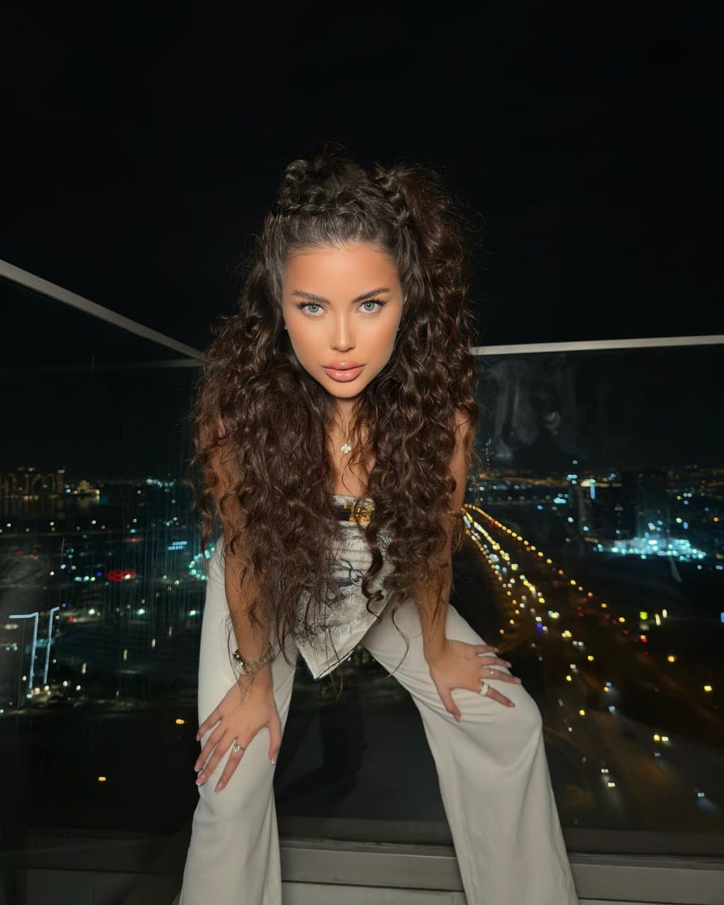 Woman in a patterned top and wide-leg trousers leans forward with hands on thighs, standing before a nighttime cityscape through a glass railing.