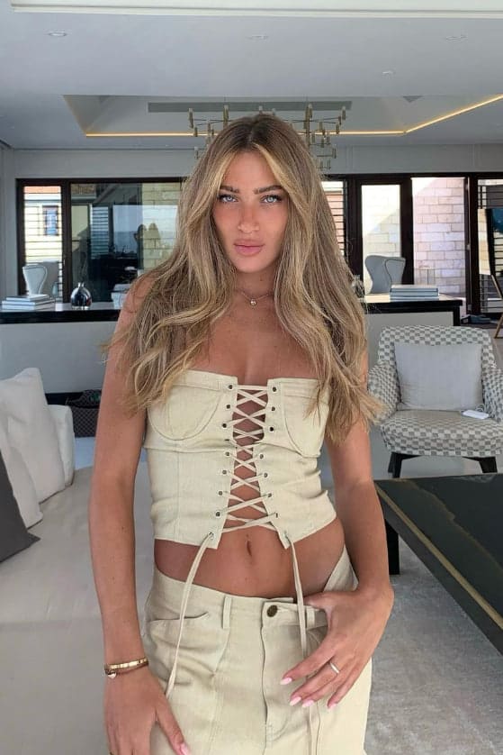 A woman stands in a living room wearing a cream lace-up corset top and matching high-waisted trousers.