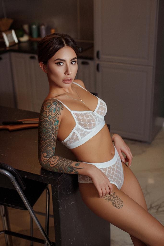 Alisha sits on a dark kitchen counter in white lace lingerie, showing a floral arm tattoo and pineapple thigh tattoo against grey cabinets.